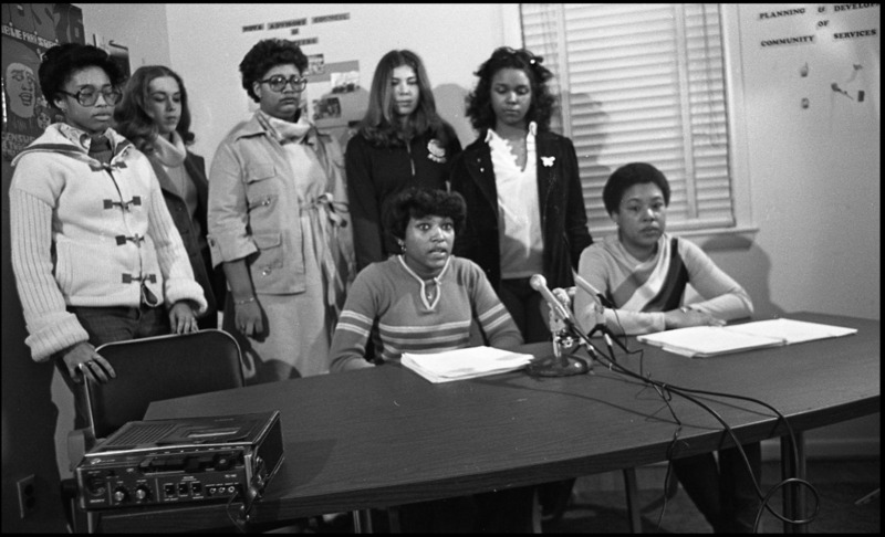 A group of white and Black students sitting and standing around a table with a microphone. Seemingly in a classroom.