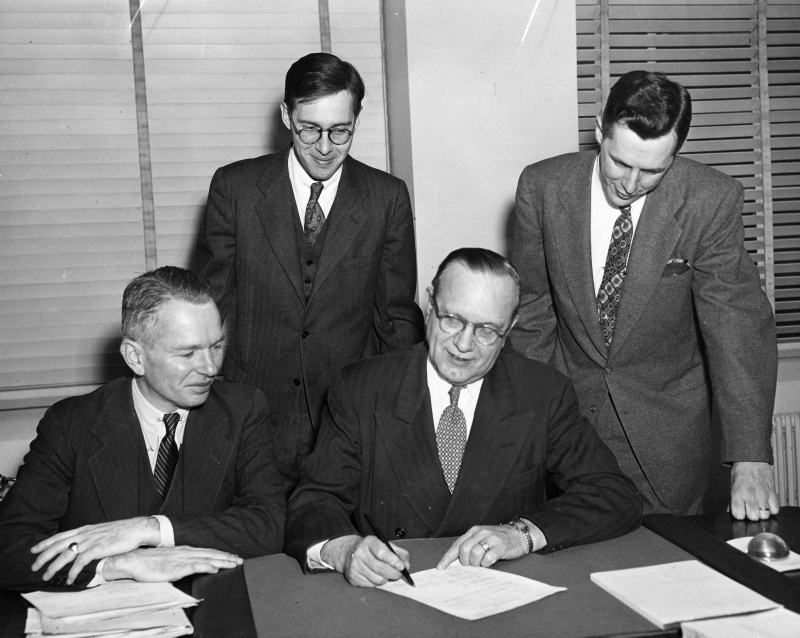 Photograph of First Signing of Reservations Under Title I of the Housing Act of 1949. Seated, from left to right: Nathaniel S. Keith, Director of the Division of Slum Clearance and Urban Redevelopment, Housing and Home Finance Agency; and Raymond M. Foley, Administrator of the Housing and Home Finance Agency. Standing, left to right: Wallace Irwin of United States Senator Howard Smith's office (New Jersey) and Walter Stults of United States Senator Robert C. Hendrickson's office (New Jersey).