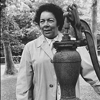 Marguerite Mott stands with outdoor water pump in 1983